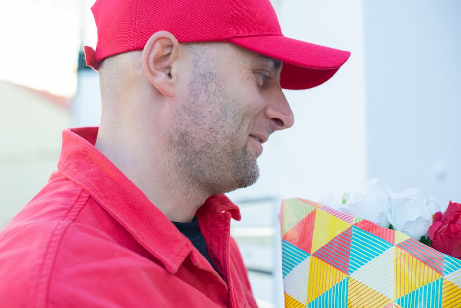 A man wearing a red cap and matching red jacket is holding a floral bouquet wrapped in colorful geometric patterned paper. The bouquet contains white and red flowers, possibly roses or peonies, with g