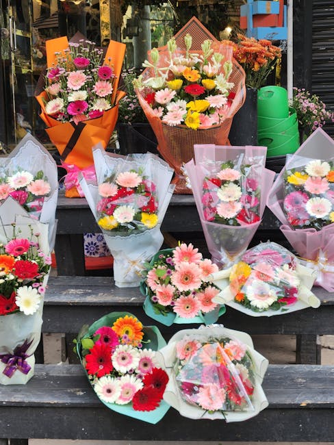 A display of various floral bouquets arranged on a dark wooden outdoor table, showcasing a range of colorful fresh flowers including pink, white, red, yellow, and purple blooms. Some bouquets are wrap