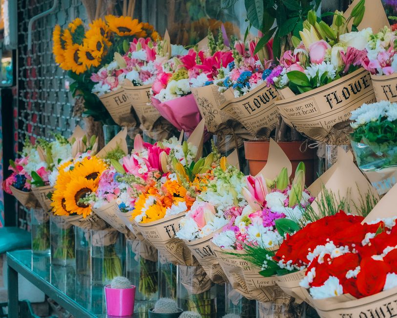 A display of fresh floral bouquets arranged in brown paper wrapping with vintage-style newspaper patterns, featuring a variety of colorful flowers including yellow sunflowers with dark centers, pink l
