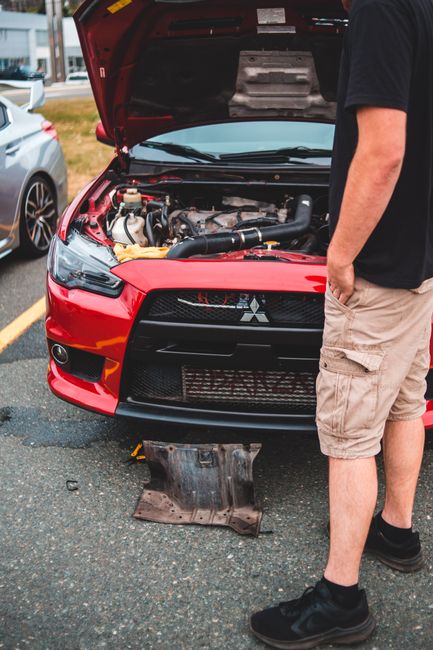 This image features a person with khaki shorts and black sneakers standing next to a red Mitsubishi car with its hood open, revealing the engine. The scene is set in a parking lot with other vehicles 