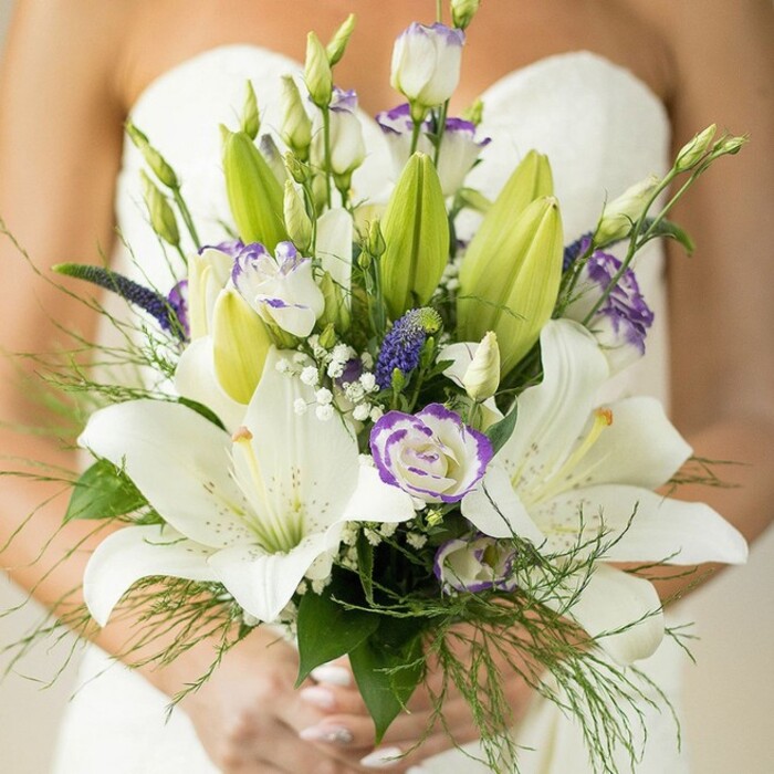 Captured in exquisite detail, this bridal bouquet is a luminous display of floral artistry, perfectly suited for Cranford's elegant wedding venues just steps from Bath Road. The arrangement is a captivating fusion of crisp white Oriental lilies, their petals fully open with touches of golden-yellow stamens and delicate freckling, complemented by unfolding buds in soft green and white. Accenting these blooms, clusters of white lisianthus grace the arrangement, their finely ruffled petals edged in understated lavender, reminiscent of early spring blossoms along the River Crane. Deeper purple highlights arise from spiky clusters of Muscari or Veronica, lending vibrant notes that echo the wildflowers found in Cranford's local meadows. Interlaced through the bouquet, baby's breath sprinkles lightness, softening the overall effect into a gentle, romantic mist. Rich green foliage-including broad, sturdy lily leaves and airy asparagus fern-frames the ensemble, their textures and hues bringing organic depth. The bride's manicured hands, adorned in a hint of pale polish, gently cup the stems, adding a human touch of grace and anticipation. With a softly blurred background of a classic white gown and bare shoulder, this arrangement, available from Cranford Flowers, is the epitome of romance and sophistication-ideal for those seeking locally handcrafted wedding florals blending traditional elegance with a whisper of contemporary artistry.