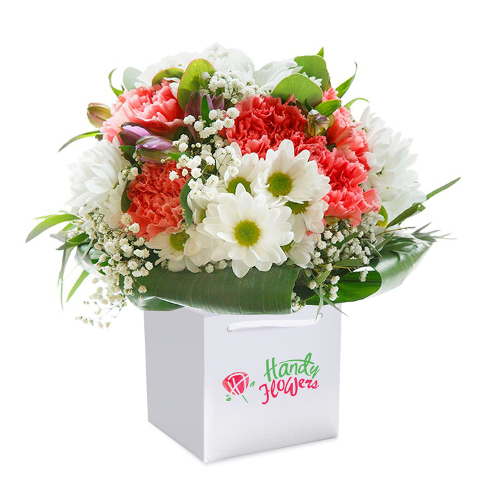 Carefully crafted by a talented Cranford flower artisan, this floral arrangement stands tall in a crisp, square white bag-every detail shining in the gentle clarity of day. The heart of the bouquet teems with soft coral and peach carnations, each frilly-edged petal forming clusters that radiate both vibrancy and tenderness. Daisies bring in notes of pure white, each crowned with a spirited yellow-green centre, embodying the cheerful essence of Cranford's green spaces. Baby's breath weaves a gossamer network among the larger blooms, lending a dreamy, weightless quality that softens the entire display. Subtle purple-tinted buds, possibly young alstroemeria, peek shyly from the left, their colour lending depth and a subtle promise of blooms to come. All these blossoms rest within a picturesque cradle of dark green, glossy leaves which fan elegantly around the base-much like the trees lining Cranford High Street or along the edge of the River Crane. The white gift bag, with its smooth satin ribbon handles, underscores a taste for minimalist delivery, making it easy to surprise a friend at Waterside Park or present as a centrepiece for a Cranford family celebration. The skillful composition, with rounded fullness and expert flower placement, assures each recipient a sense of indulgence and genuine care. This is floristry at its most heartfelt-a bouquet designed to mark birthdays, anniversaries, and new beginnings while truly reflecting Cranford's welcoming spirit.