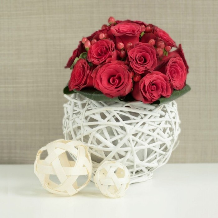 At the heart of this image is a compact, meticulously arranged bouquet of rich red roses, shaped into a dense, rounded dome that seems to hover like a soft cloud of colour. The roses, in shades of deep crimson and ruby, are packed closely together so that their overlapping petals create a continuous field of velvety texture, from the tight central buds to the slightly more open flowers around the edges. Scattered between the blooms are clusters of small, glossy Hypericum berries in a similar red tone, catching the light just enough to introduce tiny flashes of shine that break up the expanse of petals. A discreet layer of dark green leaves nestles beneath the bouquet, barely visible yet clearly supporting the flowers and giving the dome a neat, finished edge.
This elegant composition is set in a distinctive spherical vase crafted from white-painted twigs, woven into a delicate lattice reminiscent of contemporary wicker art. The criss-crossing branches form an airy globe, leaving pockets of negative space that make the vase feel light and modern rather than heavy. It rests on a smooth, bright white tabletop that reflects some of the colours above, further enhancing the clean, minimalist aesthetic. To the left of the main arrangement, two decorative balls made from pale, natural wood strips echo the vase's open weave, their neutral beige tones adding warmth and a whisper of rustic charm. The backdrop is a softly blurred wall in a light beige or taupe shade, possibly a textured fabric or wallpaper, which provides a calm, understated setting. The overall feel is of a sophisticated wedding or anniversary table in Cranford, perhaps laid out for guests after a ceremony at a nearby church or hotel, and it conveys the thoughtful, detail-focused styling that local Cranford florists bring to special gatherings and intimate home celebrations.