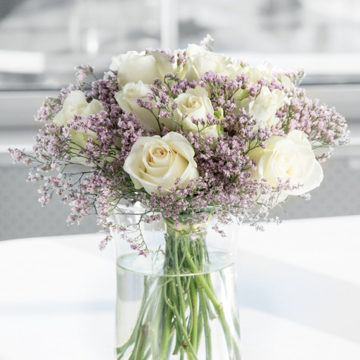 A serene floral arrangement fills the frame: a gathered bouquet of white roses and airy lavender filler flowers, presented in a tall, clear glass vase that allows every stem and detail to be seen. The roses are the heart of the design, each one open and generous, with petal upon petal spiralling inward in neat, classic formations. Their colour is a soft, creamy white with hints of ivory shadows where the petals overlap, giving them depth and a sense of quiet luxury rather than stark brightness. Between the roses, slender sprays of tiny, cloudlike flowers in pale lavender and blush-pink weave gently in and out, adding fine texture and a pastel haze that wraps the blooms in softness. These filler flowers form a light, feathery halo that makes the roses appear to float, while their cool tones bring out the subtle warmth of the white petals. The stems of both roses and fillers are clearly visible through the cylindrical vase, their green lines extending into clear, fresh water that catches small glints of light. The bouquet rests on a smooth white surface that reflects a faint glow, suggesting a tidy console or side table in a calm Cranford home, perhaps overlooking the green spaces around Cranford Park. The background is blurred into pale tones of grey and white, with no distractions, so the eye stays on the blossoms and their gentle interplay of colour and shape. The overall feeling is one of composed sympathy and graceful remembrance, the kind of arrangement a local Cranford florist would send as a thoughtful tribute to a grieving family or a quiet hospital room nearby.