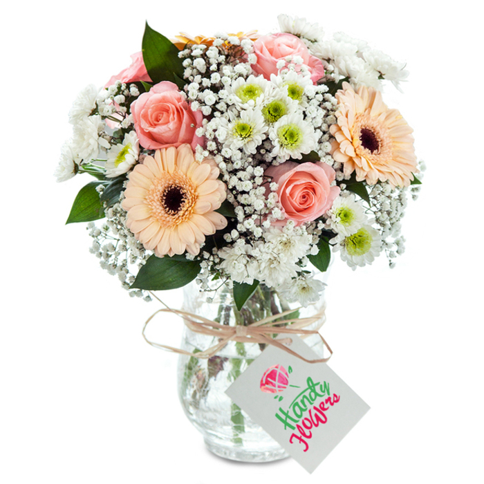An opulent bouquet of freshly cut flowers, handcrafted in Cranford and ready for local delivery, stands proudly in a clear, subtly embossed glass vase against a pure white background. Dominated by the soft glow of peach-pink roses, their generous blooms unfurl in classic spirals-a symbol of romance and hope. Two radiant gerbera daisies in hues of peach and orange inject a gentle vibrancy, highlighted by velvety brown centers that balance playfulness with elegance. Clusters of tiny white baby's breath seem to hover between each flower, bestowing an ethereal, cloud-like quality reminiscent of Cranford's quiet, open skies. Bright white chrysanthemums, their lime green hearts vivid and youthful, contribute volume and a crisp sense of renewal, while interspersed eucalyptus greens recall the neighbourhood's ancient trees. Stems are visible through the mounting vase, each one meticulously trimmed and arranged, glistening in the water to affirm their garden-fresh origin. At the neck, a band of natural raffia is tied snugly, bearing a blank white tag-inviting a personalised note to celebrate or comfort a loved one. Lit naturally and styled for beauty, this bouquet embodies the spirit of Cranford's community, poised to enhance special occasions-from anniversaries and birthdays to everyday expressions of care-with natural grace and enduring freshness.