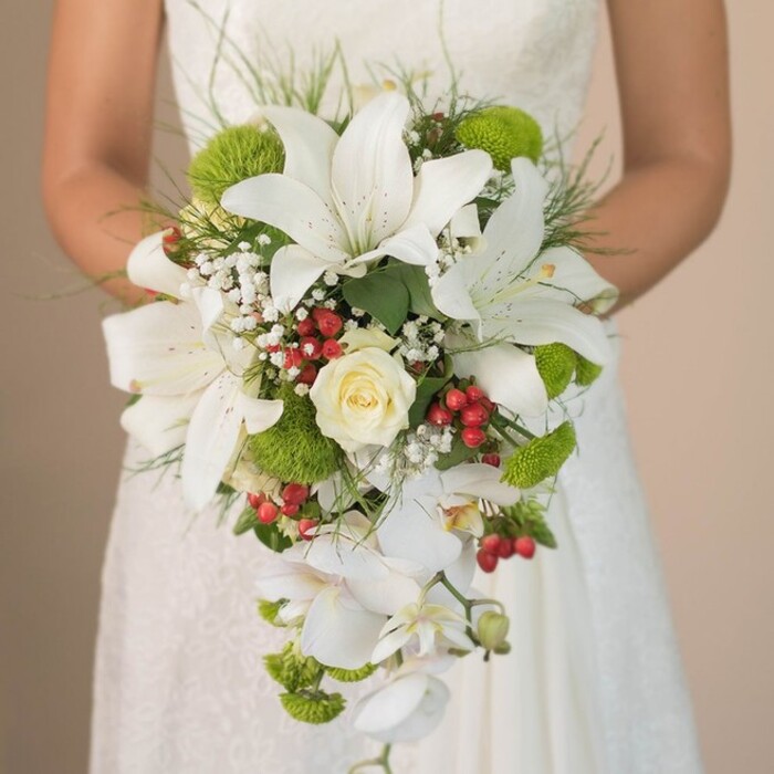 Captured in a luminous close-up that recalls Cranford's tranquil charm, this scene features a bride in a delicately patterned ivory lace gown, her arms encircling a truly showstopping cascading bouquet. The floral design is a harmonious medley: large, flawless white lilies dominate with swooping petals, each dusted with warm cinnamon freckles that catch the light. At the heart lies a resplendent, creamy white rose, its petals tightly curled in an expression of deep romance. Trailing elegantly downwards, pristine white Phalaenopsis orchids provide a fluid, waterfall-like effect, enhancing the bouquet's dramatic teardrop shape. Between the central blooms, soft tufts of baby's breath add ethereal, cloud-like softness, their star-shaped florets pooling delicately around the larger flowers. Vivid lime-green cushion mums-reminiscent of the vibrant flowerbeds in Cranford's pastures-bring playful colour and touchable texture, offset by glossy clusters of bright red hypericum berries for a joyful, contemporary twist. Lush green leaves and fine, wispy blades form an organic, natural frame, giving the bouquet an unstructured elegance, as if inspired by a walk along Cranford's leafy lanes. The bride's hands, calm but slightly trembling with excitement, are gently cradling the arrangement against the dreamy, blurred background of her gown. Altogether, this bouquet is a celebration of classic style and local freshness, perfectly suited for spring and summer weddings in Cranford and expertly crafted by Cranford Flowers for lasting beauty and charm.