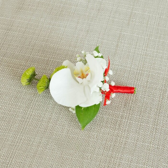 An exquisite boutonniere is carefully laid on a softly lit, light beige linen fabric, its gentle texture and natural weave creating a calm, neutral stage much like the understated décor often seen at Cranford registry office ceremonies or local hotel receptions. At the center of the composition sits a flawless white Phalaenopsis orchid, its broad, velvety petals opening in an elegant arc that suggests both strength and fragility. The orchid's throat glows with a gradient of soft yellow and subtle reddish-orange markings, drawing attention inward and giving the bloom a sense of quiet complexity, as though it holds a story within its center. Surrounding the orchid, airy clusters of white baby's breath form delicate, cloud-like sprays, their numerous tiny blossoms catching the light like miniature stars and lending a gentle, romantic haze to the design. To one side, two compact lime-green chrysanthemum pom-poms introduce a vivid, contemporary accent; their fine, spiky petals create a playful texture that contrasts beautifully with the smooth orchid and evokes the fresh, vibrant greens of Cranford's riverside paths. Nestled beneath the flowers, a large, deep green leaf acts as a grounding backdrop, while a smaller, darker leaf near the ribboned stems adds layered depth and shadow. The stems themselves are expertly bound with a rich crimson red satin ribbon, wrapped snugly and evenly before being finished with a small, precise bow or neat knot that speaks of practiced florist craftsmanship. That bold ribbon injects a confident splash of colour, making the piece perfect for a groom, usher, or formal guest seeking a refined yet distinctive accessory for celebrations across Cranford and the surrounding neighbourhoods. The overall effect is one of polished elegance, intimacy, and timeless romance.