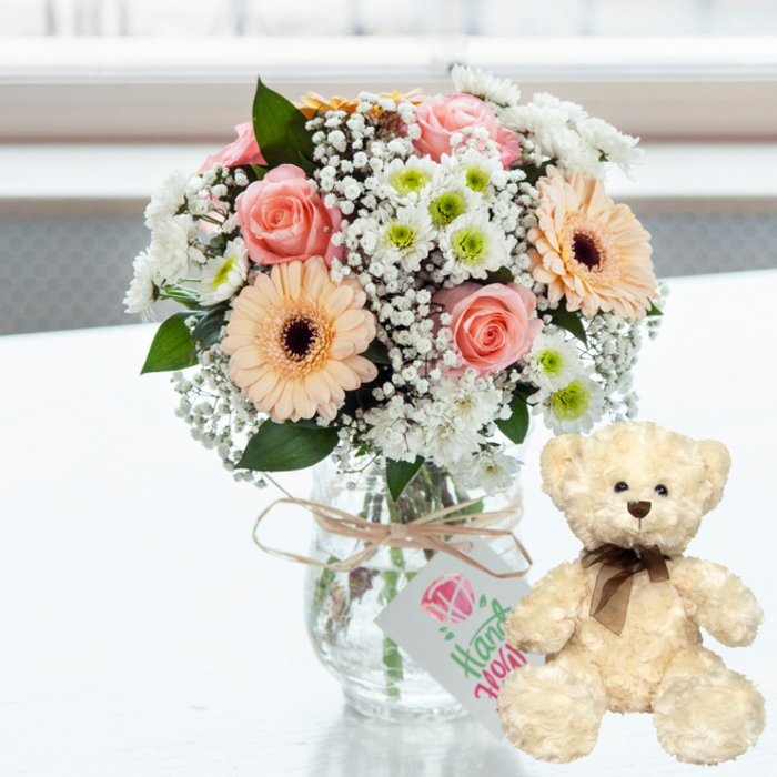 A gentle, romantic arrangement is displayed on a clean white tabletop, evoking the feeling of a carefully planned anniversary surprise in a Cranford home. At the centre of the scene stands a clear glass jar-style vase, brimming with a rounded bouquet of soft pastel flowers. Blush-pink roses, each petal curved like satin, nestle harmoniously beside peach gerbera daisies whose dark brown centres add a striking but elegant focal point. Framing these star blooms are mounds of delicate white baby's breath, their tiny blossoms giving a misty, almost cloud-like softness to the composition. Interwoven among them, small white chrysanthemums with lime-green centres bring a fresh, crisp brightness, while glimpses of dark green foliage accent the light tones and give the bouquet a natural, just-picked charm, as though sourced from gardens around Cranford. The clear vase reveals the stems gathered in the water, reinforcing the sense of freshness. Around the neck of the vase, a length of pale brown twine is tied with rustic simplicity. Dangling from it is a small rectangular white tag, printed with a pink heart and the word Handmade in graceful green script, suggesting a bespoke, heartfelt gift rather than something mass-produced. To the right, a plush cream teddy bear sits neatly on the white surface, turned slightly toward the bouquet as if admiring it. Its fur looks soft and slightly fluffy to the touch, with deep brown eyes, a rounded brown nose, and a slim brown satin ribbon tied carefully at its neck, giving it a timeless, comforting expression. The softly blurred background hints at a bright window or airy Cranford sitting room, flooding the scene with light. Together, the flowers and teddy bear create a picture of affection and reassurance, perfectly suited for celebrating anniversaries, special milestones, or simply reminding someone in Cranford that they are dearly loved.