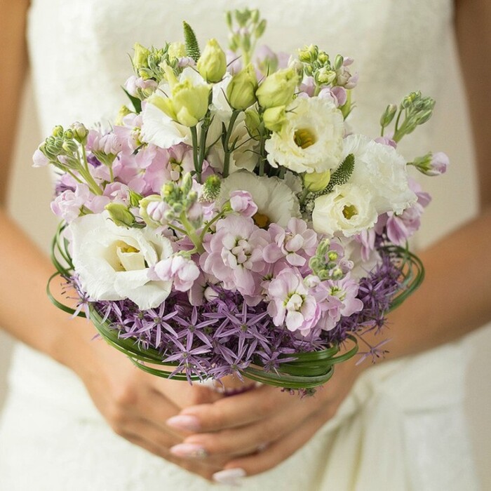 In this evocative photograph, a lush bridal bouquet is held gently in the bride's hands, her nails perfectly manicured in soft nude blush-a detail reflecting the classic taste seen across Cranford's wedding scene. The bouquet's composition brings together voluptuous Lisianthus flowers in creamy white, their frilled petals opening to reveal subtle green-yellow hearts. Pale pink and lilac stock flowers intermingle throughout, creating a layered, cloudlike effect that brings a romantic, whimsical air. Below, a ring of starburst purple Allium flowers lends striking structure: each spiky bloom a modern element, grounding the softness above with a playful yet sophisticated twist. Elegant arcs of dark green bear grass encircle the lower arrangement, carefully woven to add a sense of movement and freshness, reminiscent of the flowing meadows by the River Crane. The bouquet's stems are partially visible, wrapped with understated finesse, evidence of the florist's skilled artistry-perhaps crafted that morning at Cranford Flowers. The bride's wedding gown, seen only as a subtle white blur in the background, offers a serene, almost ethereal contrast to the colour and detail at the bouquet's heart. The fresh flowers and gentle palette conjure images of springtime in Cranford: intimate celebrations in local chapels, or outdoor vows beneath blossoming trees. Created with care, this bouquet radiates both modern urban style and timeless romance, making it an ideal choice for Cranford brides who value both local character and floral elegance.