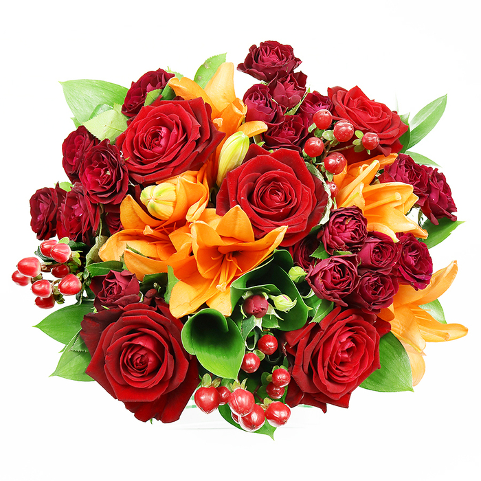 Captured from directly overhead on a spotless white background, this opulent, circular bouquet demonstrates careful floral architecture and a warm, jewel-toned palette. Several large, deep red roses dominate the centre, their plush, velveteen petals unfurling in layered spirals that suggest both classic romance and tactile richness. Bright orange Asiatic lilies are woven through the roses; some faces are fully open, revealing stamens and a lightly freckled throat, while others remain in elegant buds, lending the arrangement a sense of unfolding life and energetic contrast. Clusters of small, dark burgundy spray roses sit in close-knit groups, their miniature blooms providing concentrated colour and a luxe, textured fill between the main flowers. Glossy hypericum berries in a ruby red scatter across the bouquet like tiny, reflective jewels, catching the soft studio light and adding playful, seasonal texture. Broad green leaves - likely salal or ruscus - peek and arc around the composition, grounding the warmth with cool, verdant tones and enhancing the bouquet's fullness. The lighting is gentle and diffuse, highlighting the sheen of petals and berries without harsh shadow, and the top-down composition makes the bouquet feel both intimate and formally composed. As a local florist serving Cranford and the surrounding streets near Cranford Park and Bath Road, Cranford Flowers often recommends this arrangement for anniversaries, milestone celebrations, or as a striking centrepiece to grace a dining table before a flight from nearby Heathrow. The scent is imagined as rose-forward with a bright, spicy lily undertone - a bouquet crafted to communicate affection, celebration, and considered local craftsmanship.