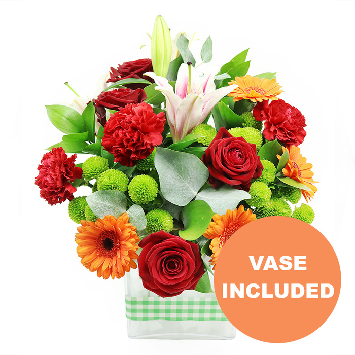 This detailed product image presents a lush mixed bouquet bursting with colour, carefully arranged in a clear, modern rectangular glass vase dressed with a fresh light green and white gingham ribbon. The flowers seem to glow against the pure white background, drawing the eye first to a cluster of deep red roses near the centre. Their tightly layered, velvety petals create a sense of depth and luxury, as if crafted petal by petal by an experienced florist from Cranford. Around them, bright orange gerbera daisies open wide, each bloom featuring a dark brown or nearly black centre that anchors the vivid petals and adds striking contrast. Threaded among these are ruffled red-orange carnations, adding frill and texture, almost like the playful bustle of a local Cranford market on a busy morning. Rising above the main body of the arrangement, several white oriental lilies provide height and elegance: some lilies are fully unfurled, revealing soft pink veining inside their trumpet-shaped petals and pollen-tipped stamens, while others remain in smooth, pale green buds that hint at blooms yet to open. Throughout the bouquet, clusters of bright lime-green pompom chrysanthemums appear like tiny spheres of colour, providing a fresh, contemporary twist and echoing the green tones of the ribbon and foliage. Deep green leaves and sprigs of silvery eucalyptus weave through the stems, adding structure, cool undertones, and a natural, almost woodland texture. In the lower right corner, overlapping the vase but not obscuring any blooms, an orange circular overlay clearly states VASE INCLUDED in bold white capital letters, signalling that this is a complete, ready-to-display gift. The overall impression is of a vibrant, expertly composed floral display, ideal for celebrations, thank-you gifts, or cheerful deliveries to homes and offices across Cranford.