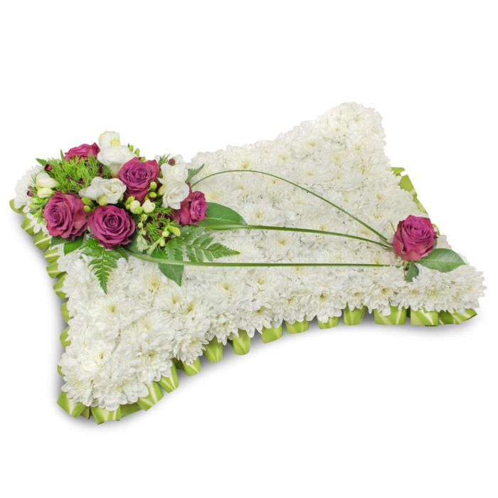 A classic sympathy pillow rendered in a serene palette, this funeral tribute is densely covered with hundreds of small white chrysanthemum blooms, creating a uniform, cushion-like surface that evokes purity and calm. The tiny florets form a tactile, almost feathery layer that invites a contemplative pause; from a short distance the pillow reads as a peaceful white plane, while close-up the individual chrysanthemum petals reveal delicate, scalloped edges. The entire cushion is edged with a light green satin ribbon folded into a continuous scallop, which gives a gentle ruffled border and a subtle note of colour without disturbing the overall quiet. Anchoring the design in the upper left is a vibrant cluster of deep fuchsia roses with velvety petals, intermingled with slender stems of white freesia and occasional cream lisianthus - these accents add contrast, fragrance, and a feeling of tender attentiveness. Fresh green foliage and feathery fern fronds fan behind and beneath the cluster, lending depth and a sense of living texture, while three curved bear-grass blades sweep from that cluster across the pillow to the opposite side, introducing an elegant, linear rhythm. A single matching fuchsia rose is placed near the lower right corner, mirroring the top grouping and completing the visual dialogue. Photographed against a clean white background with soft, even light, the tribute's shadows are minimal, focusing attention on the flowers' shapes and tones. Designed for memorials and remembrance services in Cranford - whether delivered to local churches, chapels, or family homes close to Cranford Park - this pillow communicates solace, dignity, and the quiet comfort of careful floral craftsmanship.