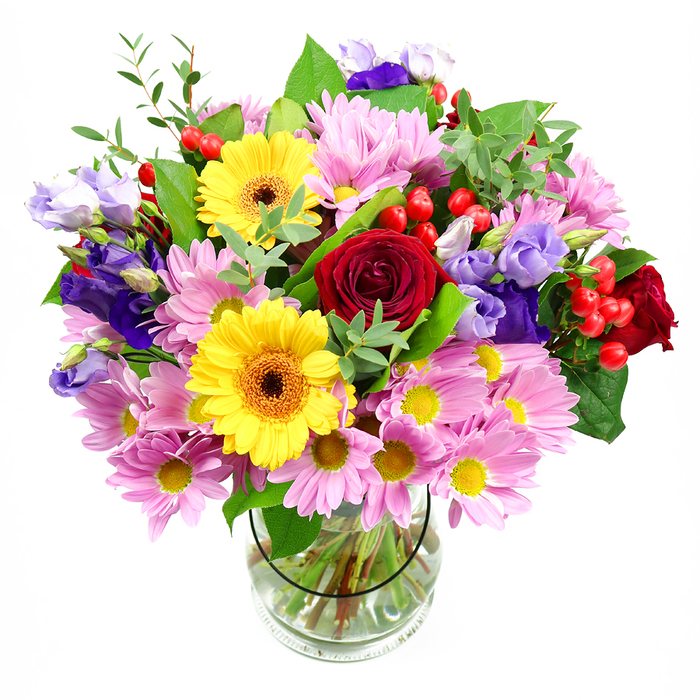 A bright, abundant bouquet rises confidently from a crystal-clear glass vase, its colours singing against a pure white background that resembles a softly lit studio in a Cranford high street shop. Dominating the centre, sumptuous red roses, richly shaded and velvety to the eye, give the arrangement a heart of classic romance. Nestled close by, two vivid yellow gerbera daisies with warm, dark centres exude cheerfulness, their wide, flat petals radiating like small suns. Encircling these focal flowers are generous sprays of pink daisies and chrysanthemums, each blossom made up of slender petals fanning out from a golden eye, adding both volume and a tender, welcoming tone. Tall stems of lisianthus emerge through the bouquet, their bell-shaped blooms shaded in lavender and purple, some still in bud, lending a layered, almost painterly effect and a note of quiet sophistication. Tucked between the petals, rounded red hypericum berries catch the light, providing bright, textural punctuation that feels playful yet refined. The arrangement is wrapped in a frame of lush greenery: glossy, deeper green leaves give weight and structure, while lighter, more delicate sprigs add movement and the suggestion of a soft breeze passing through. Through the glass vase, you can see the straight, evenly cut stems immersed in clear water, a testament to careful conditioning and attention to detail. This bouquet feels perfectly suited to life in Cranford-ideal for sending to a friend after a walk in Cranford Park, celebrating a special family moment at home, or simply turning an ordinary day into something more memorable with its mix of colour, fragrance, and thoughtful design.
