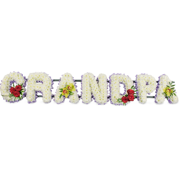 The image shows a striking funeral flower tribute spelling out the word GRANDPA in large, upright capital letters arranged horizontally, photographed against a plain white backdrop so that nothing distracts from the flowers themselves. Each letter is heavily padded with small, snowy white chrysanthemums, tightly packed to form a smooth, plush surface that gives the tribute both structure and softness. Around the outside edge of every letter runs a fine trim of soft lavender-purple ribbon, lightly ruffled, which adds a gentle touch of colour and helps each shape stand out clearly from the bright background. At the start of the word, tucked into the open curve of the G, sits a compact cluster of three or four deep red roses surrounded by fresh green foliage, creating a point of rich colour and emotion. The third letter, an A, carries a spray of yellow-green orchids, some with subtle reddish markings at their centres, their slender petals and sculpted forms lending an elegant, slightly exotic note. Mirroring the first letter, the curve of the D also holds a similar grouping of deep red roses and greenery, while the final A at the end of the tribute repeats the yellow-green orchids, visually tying the whole word together. Behind the flowers, slim horizontal bars and a dark support frame hold the letters neatly in line, ready to rest beside a coffin, at a memorial service, or by a grave in Cranford's local cemeteries or churchyards. The overall mood is calm, respectful, and deeply personal, reflecting a loving family's desire to say goodbye to their grandfather with purity, warmth, and enduring remembrance.
