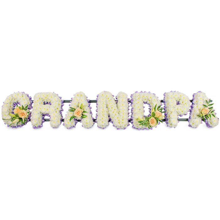 This photograph captures a refined funeral flower arrangement spelling the word GRANDPA in large, uppercase letters arranged in a straight horizontal line, as if ready to be placed at the head of a service in Cranford. Each letter is fully filled with a dense layer of white spray chrysanthemums, or similar small-petalled flowers, carefully inserted so no gaps of foam or support are visible. The blossoms create a thick, padded effect, suggesting softness to the touch and conveying a sense of purity and peace. Surrounding the edge of every letter is a narrow band of light lavender or pale purple flowers, delicately woven along the perimeter to define the shapes and add a subtle contrast that catches the eye. Throughout the tribute, carefully chosen accent clusters of flowers are positioned with restraint and elegance. The G is adorned with a small spray of peach roses, some just opening, nestled among dark green foliage and a few slim white buds, perhaps lily or similar blooms. A single peach rose with greenery rests on the first A, while the D holds another small grouping of roses and leaves, and the final A repeats the gentle touch of a single rose. These peach accents introduce warmth and a hint of tenderness against the cool whites and lilacs. All seven letters sit on a discreet dark bar, likely metal or green-painted, that provides structural support without drawing attention away from the flowers. Set against a plain white background, every colour and contour stands out clearly, evoking a calm, respectful mood-an individualised tribute for a cherished grandfather, made for moments of remembrance and quiet reflection.