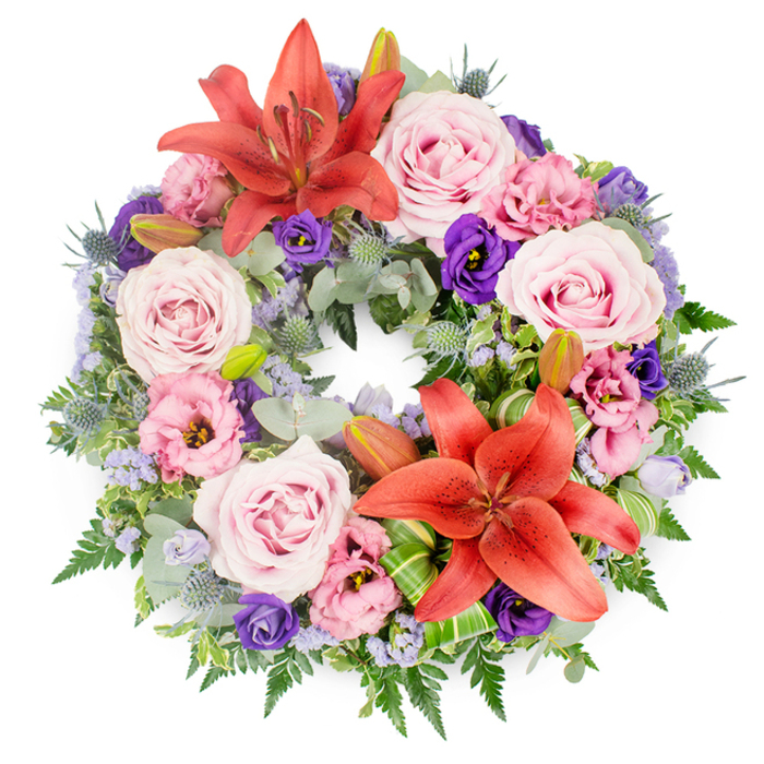 Centered on a bright white background is a meticulously crafted circular funeral wreath, its dense ring of flowers and foliage forming a complete halo of colour and remembrance. This tribute, the kind a Cranford family might choose for a service after a peaceful walk through Cranford Park, is built upon a foundation of rich greenery, with every space thoughtfully filled. Two large coral-orange Asiatic lilies serve as striking focal points, one gracing the upper curve of the wreath and another anchoring the lower section. Their broad, slightly recurved petals and clearly defined stamens bring a vibrant, sunrise-like intensity to the arrangement, standing out against the cooler and softer hues around them. Embracing these lilies are numerous roses in flattering shades of blush and lavender-pink, each rose fully open, with tightly layered petals that seem to cradle unspoken sentiments of love and farewell. Nestled between the roses are ruffled lisianthus flowers, some a pale, almost cloud-soft pink, others a deep, regal purple. Their fluted, cup-shaped blooms add movement and a sense of gentle flutter, as if stirred by a light breeze. Scattered throughout the wreath are delicate sprays of tiny lavender-blue filler flowers, likely statice or limonium, which create a subtle, misty veil that unites the stronger, more defined blooms. The foliage foundation is varied and textural: rounded, silvery-green eucalyptus leaves catch the light; darker, glossy leaves lend depth; fine fern fronds and other airy greens fill out the circle. Blue-grey eryngium thistles, with their spiky, star-like heads, punctuate the design and bring an element of quiet strength. Several tightly closed lily buds rest among the greenery, hinting at blossoms that will open in the days following the service. Near the lower lily, carefully folded and shaped bright green leaves frame the bloom, showcasing the skill of a professional Cranford florist. The overall composition exudes balance, dignity, and deep sympathy, making it a fitting wreath for funerals, memorials, or graveside tributes in the local area.