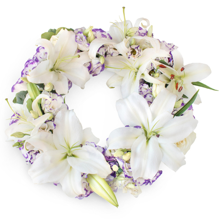 A beautifully composed circular sympathy wreath is photographed front-on against a bright, unblemished white background, allowing every flower and leaf to stand out clearly. The wreath forms a perfect, unbroken ring, filled edge to edge with lush white Oriental lilies whose long, elegant petals curve outward like folded satin. Many of the lilies are fully open, each revealing a soft green centre and upright stamens tipped with warm amber-brown anthers, small details that bring a touch of natural vibrancy to the calm palette. Between these commanding blooms, clusters of lisianthus unfold in layers of ruffled petals, white at their core and brushed with whispers of lavender and violet along their edges, lending the wreath a gentle, watercolour-like softness. Occasional lily buds, still tightly furled, and a few discrete ivory buds reminiscent of miniature roses add nuance and depth. Threads of dark and mid-green foliage, along with eucalyptus-style sprigs, nestle quietly behind the blossoms, forming a supportive base that gives the circle structure without drawing attention away from the flowers. The overall effect is tranquil and dignified, evoking the hush of a chapel or the reflective stillness of a walk along the tree-lined streets of Cranford on the way to a service. This refined lily and lisianthus wreath feels especially suited for funerals, memorials, or graveside tributes in the local area, offering a graceful way to commemorate a life with purity, tenderness, and enduring respect.