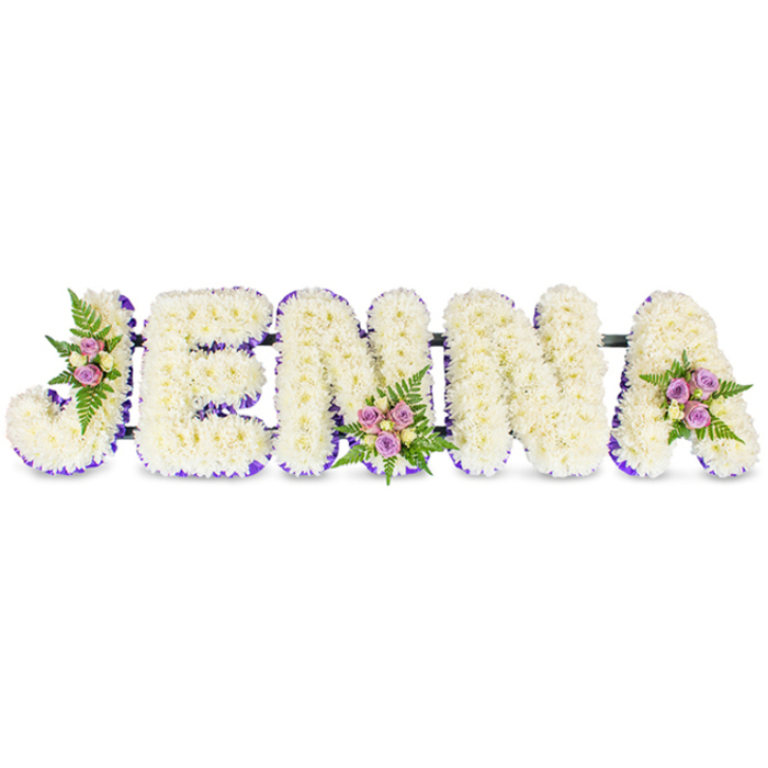 A compassionate, personalised floral display forms the name JENNA in five substantial, three-dimensional capital letters arranged horizontally against a clean white background. Each letter is lavishly built from hundreds of small, creamy white chrysanthemums or similar delicate florets, creating a soft, inviting surface reminiscent of a white blanket of petals. A slender band of vivid purple-whether a ribbon or a halo of tiny lilac blooms-runs along the inner edge of each letter, offering a refined contrast that outlines the shape and adds visual poise. The letters are subtly linked by narrow dark bars to ensure stability without drawing attention away from the floral work. Tasteful clusters decorate three of the letters: one placed on the curve of the J, a second at the base of the first N, and a third adorning the top of the A. Those clusters bring together gentle blush-pink and lavender spray roses, accented by a few soft cream or pale yellow roses and nestled into bright green fern fronds, introducing small islands of colour and a softer fragrance amid the white. The overall impression is one of peacefulness and thoughtful craftsmanship, ideal for a commemorative display at a funeral, wake or celebration of life. Locally, families from Cranford village and nearby streets often choose this style when holding services close to Cranford Park or arranging tributes for gatherings near the Bath Road; the design reads clearly from the front and carries a mood of humble reverence.