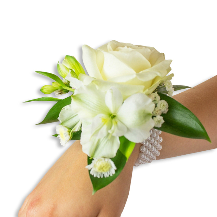 Seen from a slightly elevated angle, this wrist corsage is a composed portrait of white florals and deep greenery resting on a fair-skinned wrist against a neutral white field. The central element is a creamy ivory rose, its layered petals curling outward with a soft, velvety texture that reflects the light with a pearlescent sheen. Below and a touch to the left, a large, trumpet-like white blossom-likely an alstroemeria or a small trumpet lily-spreads its slightly ribbed petals to reveal a faint lime-green throat, a gentle focal contrast within the pale palette. Fine sprays of Gypsophila nestle around the main blooms like drifting mist, providing an airy, delicate texture, while a cluster of small white daisy-like florets with bright golden centres adds a tender, nostalgic accent close to the wrist. Sprigs of vibrant green buds punctuate the design, their taut forms promising future bloom and adding dimension. The flowers are balanced and framed by several broad, glossy dark-green leaves with a smooth, waxy finish that gives the composition a polished, garden-fresh look. A multi-strand faux-pearl bracelet secures the arrangement: rows of uniformly round pearls encircle the wrist, offering a classic, tactile elegance that complements the flowers' softness. The overall mood is serene and celebratory-ideal for a wedding, prom, or formal occasion-and evokes the gentle mornings and calm green spaces around Cranford and neighbouring Hounslow.